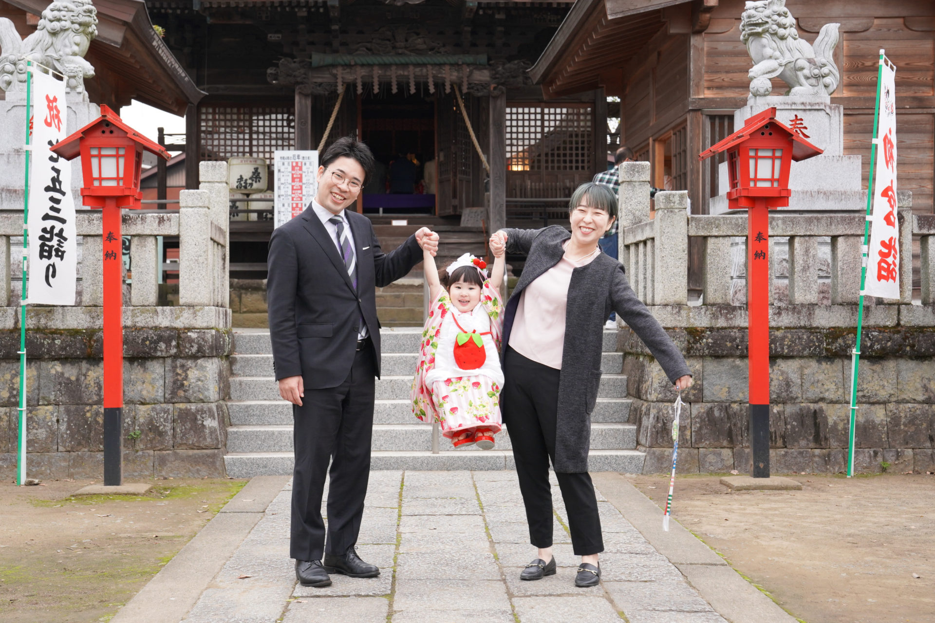神社の前で、ピンクと赤の着物を見に纏い、両親と手を繋ぎ高い高いをしてもらっている3歳の女の子の七五三