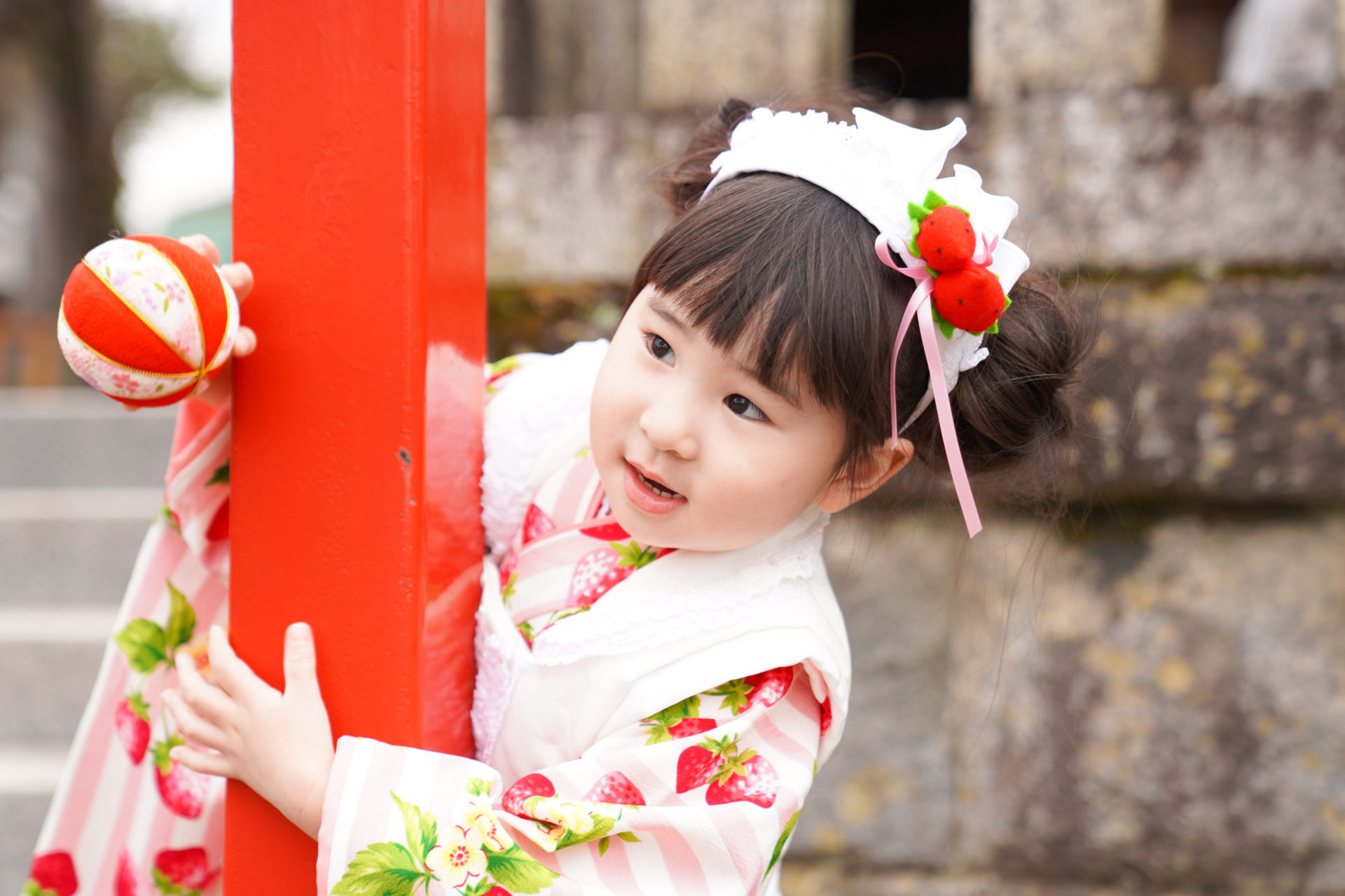 神社の境内で、ピンクと赤の着物を見に纏い、赤い柱から顔を覗かせる、3歳の女の子の七五三