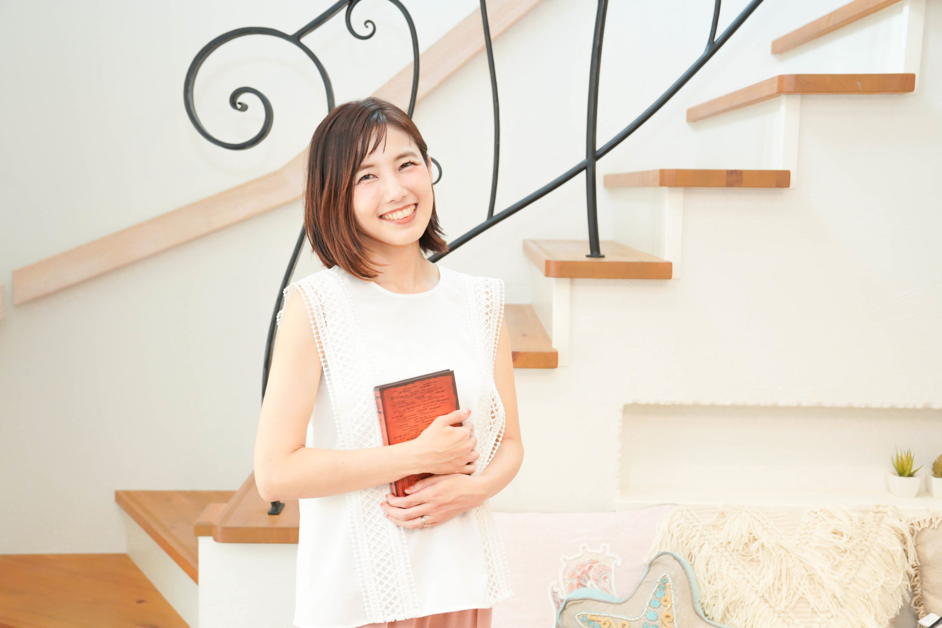 白いトップスを着て、本を持ち、階段の前で笑顔でこちらに視線を向ける女性
