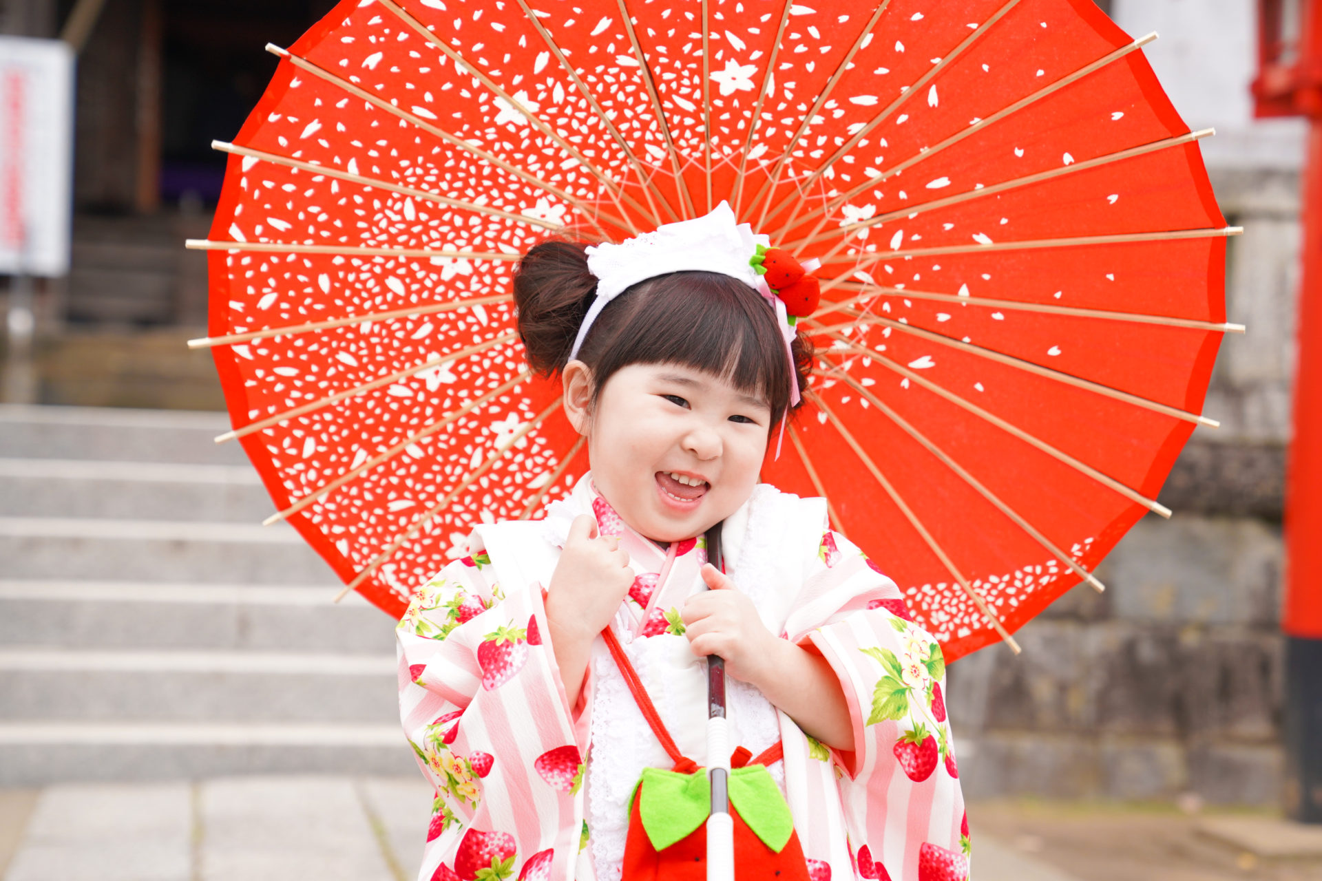 神社の境内で、ピンクと赤の着物を見に纏い、真っ赤な和傘をさし満面の笑みの3歳の女の子の七五三