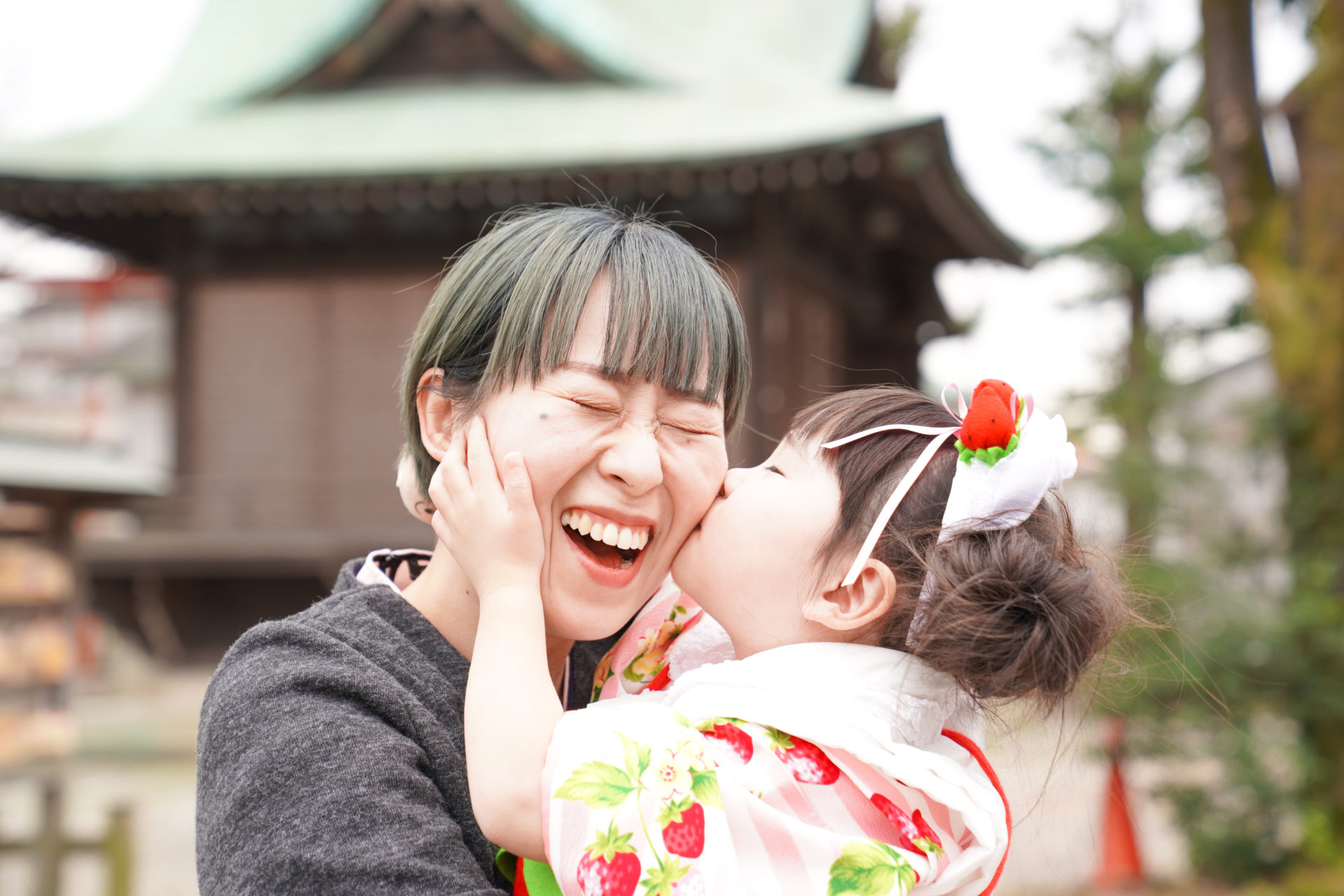神社の境内で、ピンクと赤の着物を見に纏い、ママのほっぺにキスをする、3歳の女の子の七五三